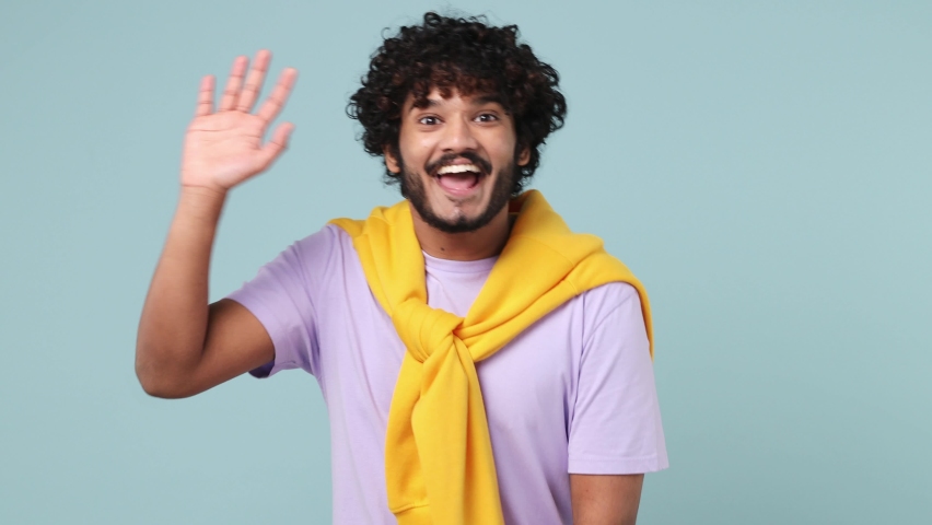 Happy cheery young bearded Indian man 20s wears violet t-shirt look around for friend find waving meet greet with hand as notices someone isolated on plain pastel light blue background studio portrait Royalty-Free Stock Footage #1088333501
