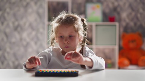 Upset little girl student plays with toy abacus learning mental arithmetics at table in kindergarten classroom close view slow motion