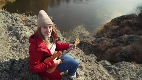Caucasian woman sits on rock and plays the ukulele. Her blonde hair is blown by the wind. She plays the strings of small guitar. Autumn, yellow leaves on trees. Hobbies, string, music