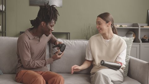 Medium slowmo shot of young Caucasian woman showing her high-tech prosthetic arm to African-American male friend then bumping fists, sitting together on sofa at home