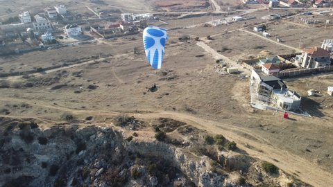 Aerial drone view of a man flying a white and blue paraglider over a hill and trees to the sea waves near the rocks. Active paraglider flight over the seascape with clear skies at sunset. Extreme sport