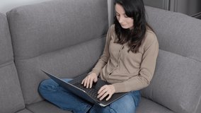 Ukrainian woman typing text on laptop keyboard while sitting on comfortable couch in living room at home. Freelance writer person doing distant work online. Brunette female networking with clients