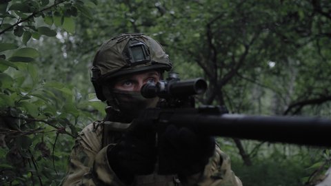 Close-up, an armed soldier commando with a sniper rifle, in a dense forest, protects the front line, in combat readiness aims at the enemy through an optical sight. Equipped soldier in action.
