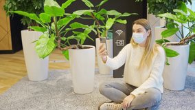 a young woman in a protective mask sits on the floor near home green plants and a wi-fi sign and talks on a smartphone via video link