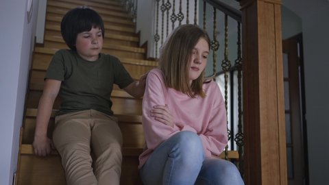 Irritated teenage girl pushing away hand of little boy leaving as sibling sitting on stairs hugging knees. Portrait of argued Caucasian sister and brother at home indoors. Rivalry and lifestyle