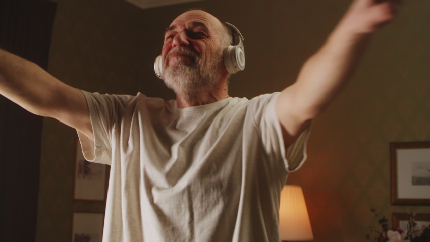 Low angle of happy elderly male in white t shirt and wireless headphones, listening to music and swinging arms during fitness training in morning at home
