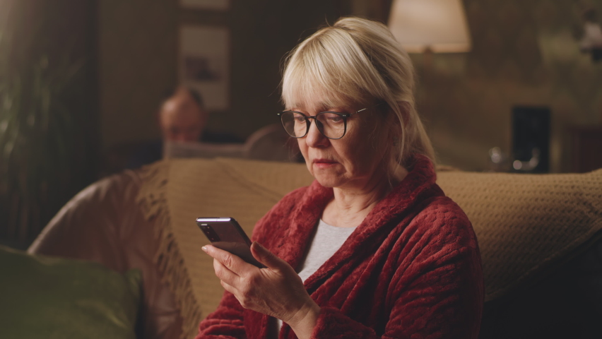 Shocked elderly female in glasses looking around after reading terrifying news on mobile phone while sitting on sofa in living room at home | Shutterstock HD Video #1088363645