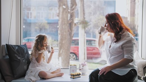 tea party. mother and little daughter are drinking tea with sweets, sitting by the window, on a large windowsill.