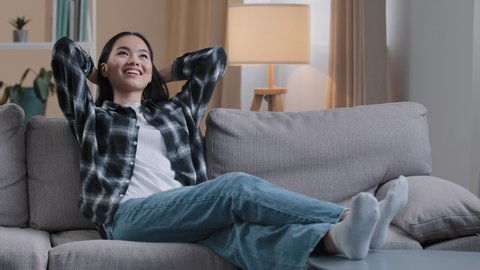 Calm happy tired asian woman housewife resting on soft couch holding hands behind head korean lady relaxing on comfortable sofa napping feel stress free at home lounge peaceful resting indoors cozy