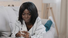 Young african american woman lying on couch resting relaxing chatting on video call using smartphone looking at phone screen waving hello talking on webcam via video conference friendly conversation