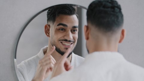 Funny arabian indian bearded man speaking to reflection looking in bathroom mirror. Smiling sexy millennial guy narcissist speaks to himself rehearses speech performance oratory affirmation in morning