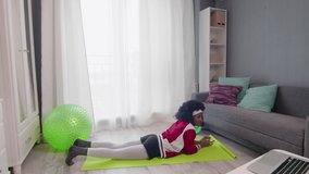 Young african american woman hippy in colourful sportswear with curly afro hairs doing plank and watching fitness video tutorial online on laptop computer