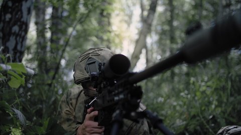 An armed special forces soldier with a sniper rifle sits in ambush in a dense deciduous forest, defending the front line during a special operation. Soldier in full gear