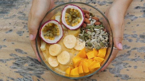 Delicious raw vegan smoothie bowl with exotic tropical fruits. Female hands puts organic breakfast on wooden table view from above. High quality 4K super food concept footage. Thailand.
