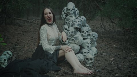 Fantasy video with noise. gothic woman vampire sits near mountain of skulls victims, bares predatory teeth, hisses into camera. Wild angry hungry face long fake fangs. Halloween costume, decorations