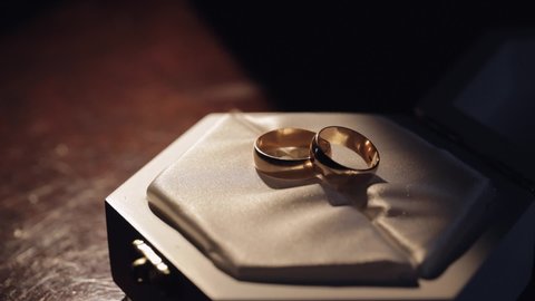 Two gold wedding rings lying on white gift box shining with warm light close up macro. Pair of rings on dark background. Gifts and romance, decor of bride and groom. Matrimony event. Slow motion
