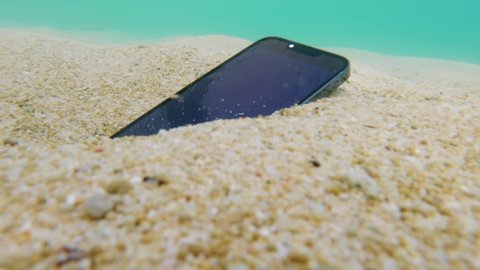 Close-up live camera footage of beautiful underwater sea bottom with white sand swirling with stream and cellphone with blue screen for chroma key being taken by anonymous man. Waterproof handphone.