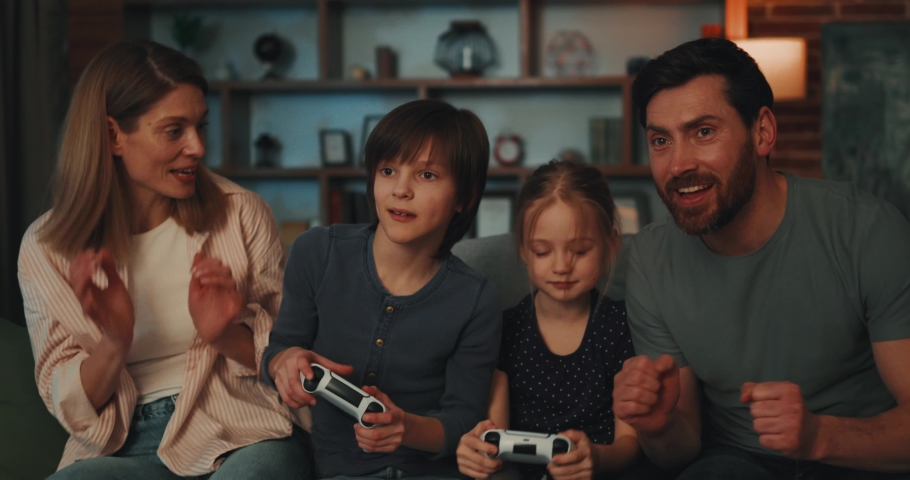 Portrait of Young Mum and Dad Cheering for Children Playing Video Games at Home. Joyful Girl and Boy Competing sitting on Sofa using PlayStation holding Joystick. Girl Sister Winner. Family Team | Shutterstock HD Video #1088386565