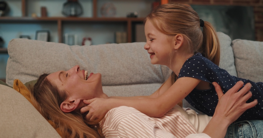 Carefree Mother and Cute Small Child Daughter  laughing lying on couch. Happy  Womn, enjoying of motherhood, hugging Little Girl Spending Time Together at Home 