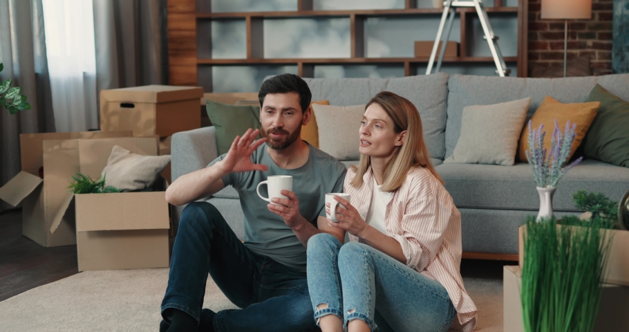  Loving Couple Drinking Coffee on the Carpet in Spacious New Home. Loving Man and Woman talking and resting after Moving to Apartment sitting near Boxes and Sofa Royalty-Free Stock Footage #1088386667