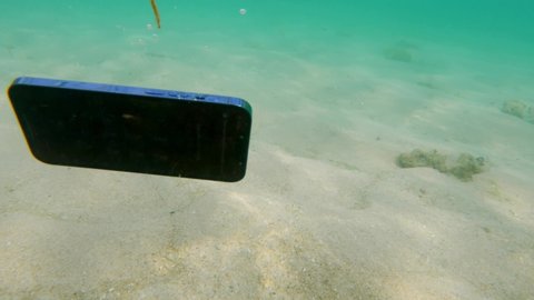 Unstable live camera shot of modern smartphone sinking under ocean water and diving into white sand with texture of sun dogs. Waterproof mobile phone. A modern smartphone is drowning in the ocean. 