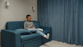 Preteen black boy holding joystick, sitting on couch. Mom approaching son, holding laundry hamper indoors. Housewife busy with chores. Kid obsessed with video game, ignoring mother's request at home