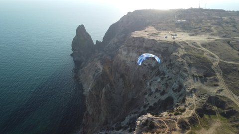 Aerial drone view of a man flying a white and blue paraglider over a hill and trees to the sea waves near the rocks. Active paraglider flight over the seascape with clear skies at suset. Extreme sport