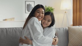 Sincere family love. Zoom in portrait of happy asian mother and little daughter embracing at home, sitting in bathrobes on sofa and smiling to camera, slow motion - Powered by Shutterstock - Get 15% off with code: PIKWIZARD15