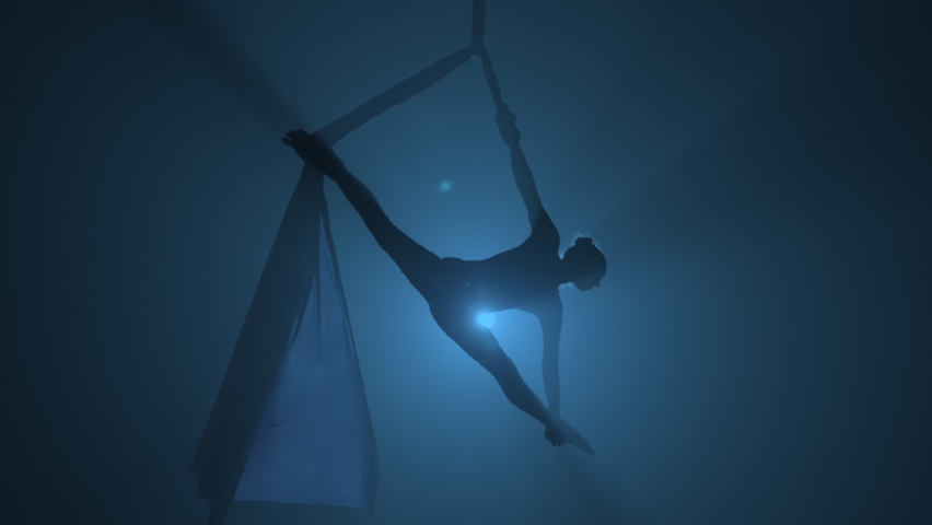 Silhouette of graceful aerial gymnast performing acrobatic stunts and stretching on aerial silk. A young woman performs with circus show at a height against smoky dark backlit background. Slow motion. Royalty-Free Stock Footage #1088393937
