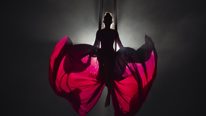 Silhouette of an aerial circus gymnast performing on aerial silk and waving canvas like wings. Beautiful circus show performed by young woman acrobat on black background with backlight. Slow motion. Royalty-Free Stock Footage #1088393951
