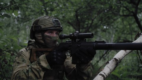 Close-up, an armed soldier with a sniper rifle, in a dense deciduous forest, protects the front line, in combat readiness aims at the enemy through an optical sight. Equipped soldier in action