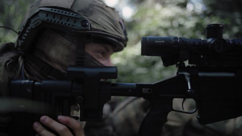 Close-up, an armed special forces soldier with a sniper rifle sits in ambush in a dense deciduous forest, defending the front line in a special operation. Soldier looking through a scope