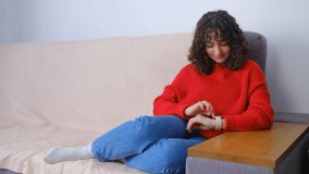 Young female browsing smart watches on couch at home. Beautiful white woman with curly dark hair using modern pink wristwatches for communication online during lockdown. Pretty brunette use gadget