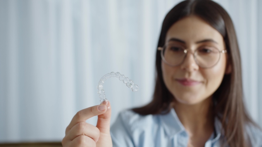 Happy Attractive Woman Smiles And Sitting In Cozy House And Holding Invisalign Braces. Putting On Transparent Plastic Retainers Or Tooth Whitening System. Concept Of Dental Healthcare And Orthodontic | Shutterstock HD Video #1088395983
