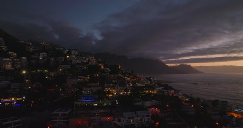 Luxury residences and apartments in slope above water. Forwards fly above sea coast in town at dusk. Cape Town, South Africa