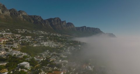 Forwards fly along fog rising from sea along coast. Residential houses in slope under rocky escarpment. Cape Town, South Africa