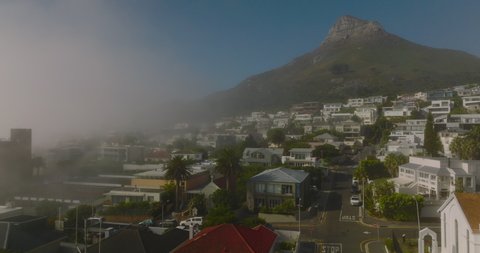 Fly above houses in residential borough of touristic destination. High rocky peak ahead. Morning fog rising from sea. Cape Town, South Africa