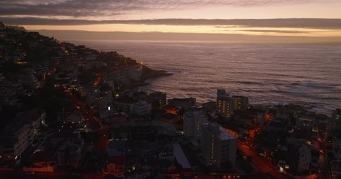 Fly above buildings in urban neighbourhood at sea coast at dusk. Rippled water surface and colourful sky. Cape Town, South Africa