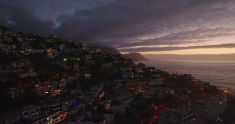 Forwards fly above residential buildings in town. Aerial view of sea coast at sunset. Cape Town, South Africa