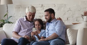 Older man, little cute grandson and young adult millennial son sit on sofa having fun using smartphone. Three generations men, multi generational family spend leisure on internet, modern tech concept