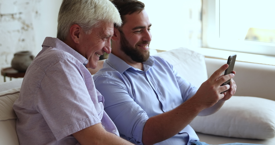 Close up smiling older man and adult 35s son sit on sofa with smartphone, have fun, enjoy new mobile application looking happy spend time together on weekend at home. Leisure using modern tech concept Royalty-Free Stock Footage #1088401253