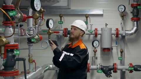 The engineer checks the position of the valves on the pipeline and the readings of the pressure gauges on the smartphone. A bearded man in a white helmet at work in heavy industry.