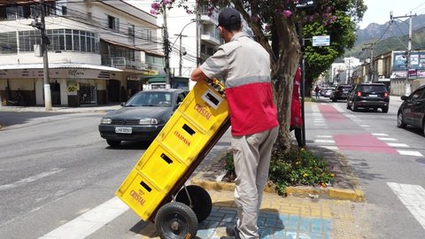 Teresopolis, RJ, Brazil - March 18th 2022 - Beer delivery man. Man carrying cases of beer on a cart. Yellow beer boxes from Grupo Petropolis. Deliveryman crossing the street with several beer bottles.