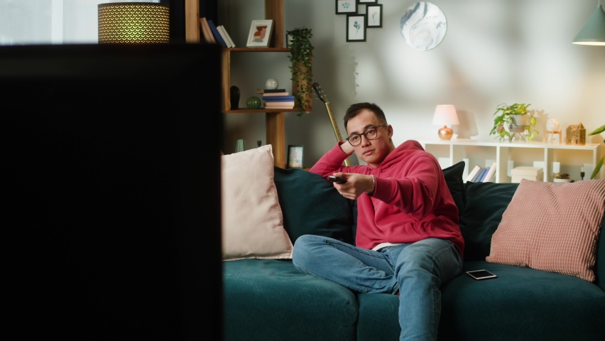 Asian man watching television. Young Korean guy changing channels on tv, using remote control, sitting on sofa in living room, male person relaxing at home, watch film.