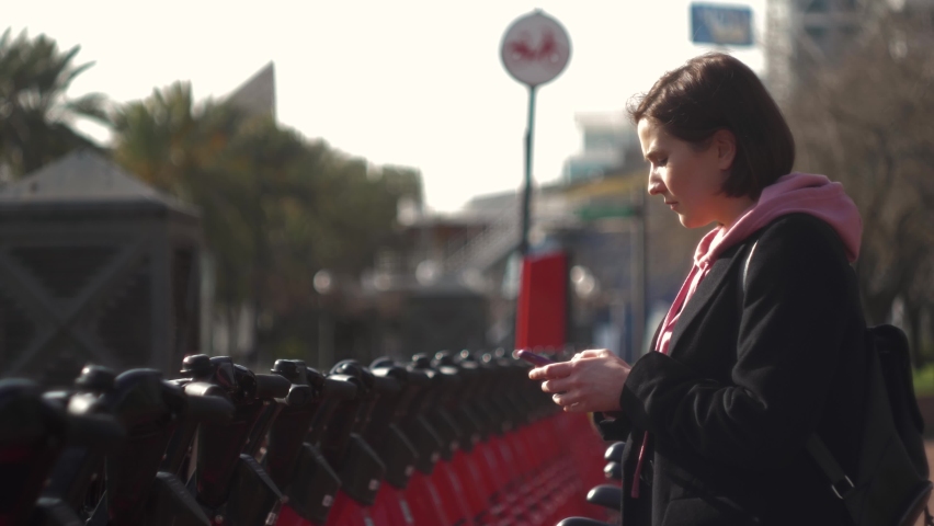 Tourist woman take Electric Kick scooter or bike bicycle in sharing parking lot, at night phone application. New sharing business project started in city, eco transportation