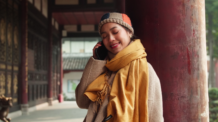The beautiful girl,talking on cellphone,standing under the Chinese traditional wooden structure buildings, with a smile on her face, wearing a yellow scarf and wool hairband,