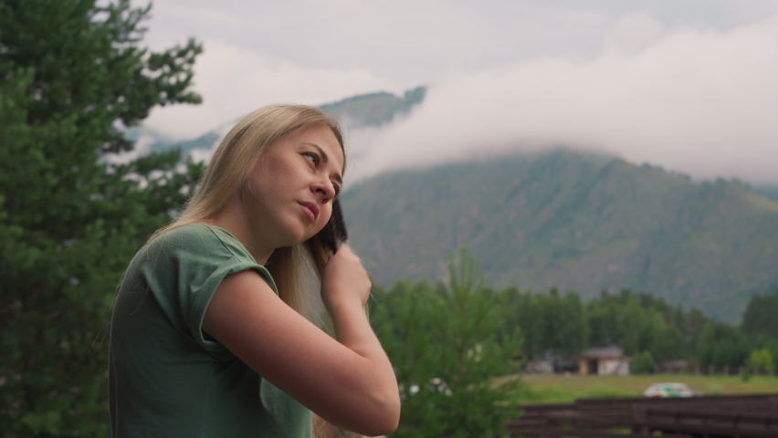 Attractive young woman brushes long hair standing against old mountains covered with descending white misty clouds at resort slow motion