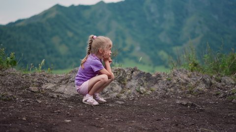 Tired little girl sits on haunches leaning head on palm on rocky hill against distant mountains at national reserve on nasty day slow motion