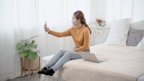 Asian woman sitting on bed taking selfie while using laptop working. Happy female making video call relatives or bestfriend with smartphone at home. Concept of technology.