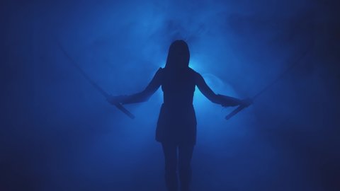 Dark black silhouette fantasy woman warrior assassin holding two katana swords. Black background blue neon light full smoke. Fairy soldier girl hand raised. Armed sexy girl princess. Medieval weapon